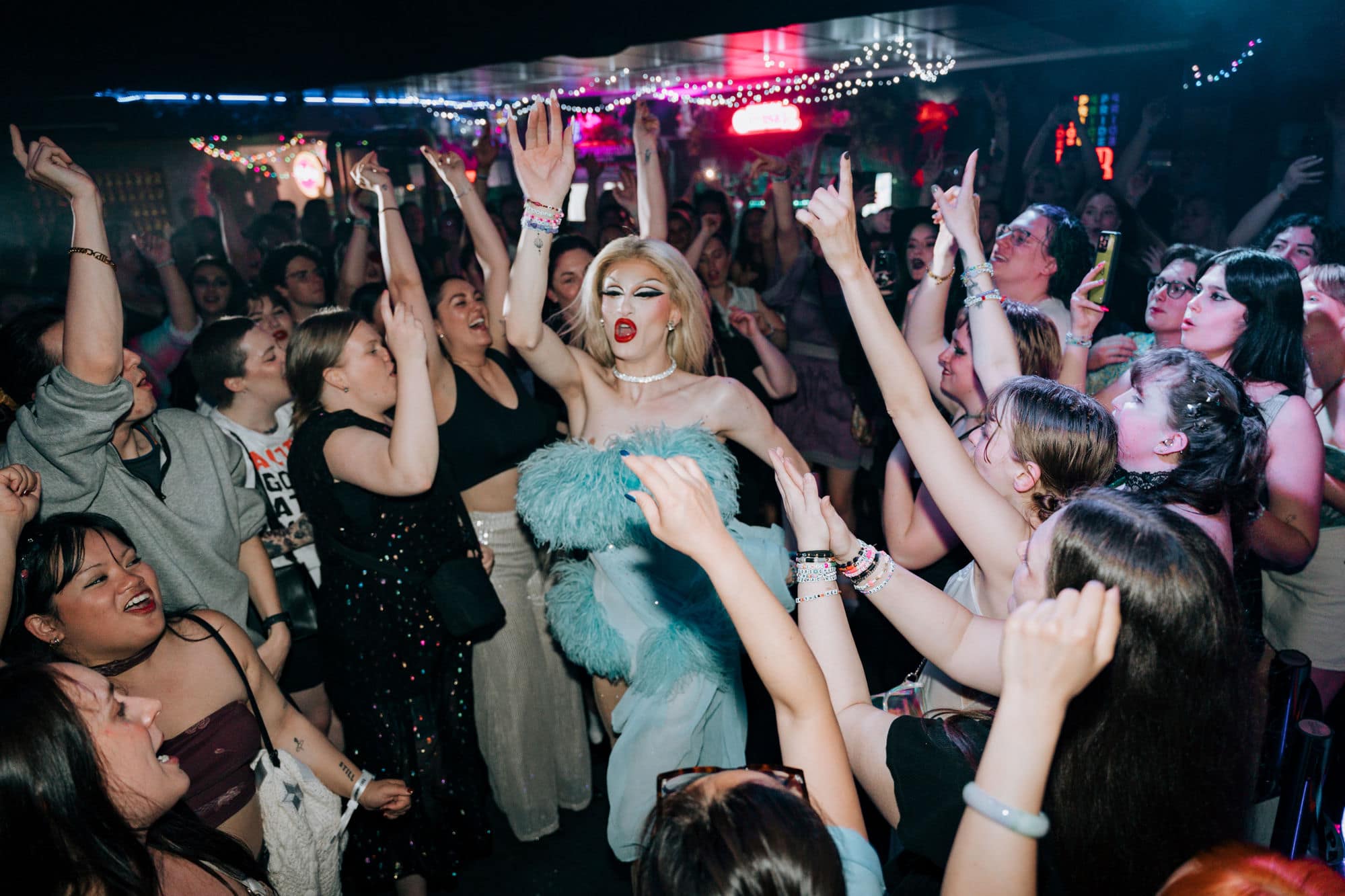 A drag performer and a packed crowd with hands in the air on the dancefloor at Pride of our Footscray
